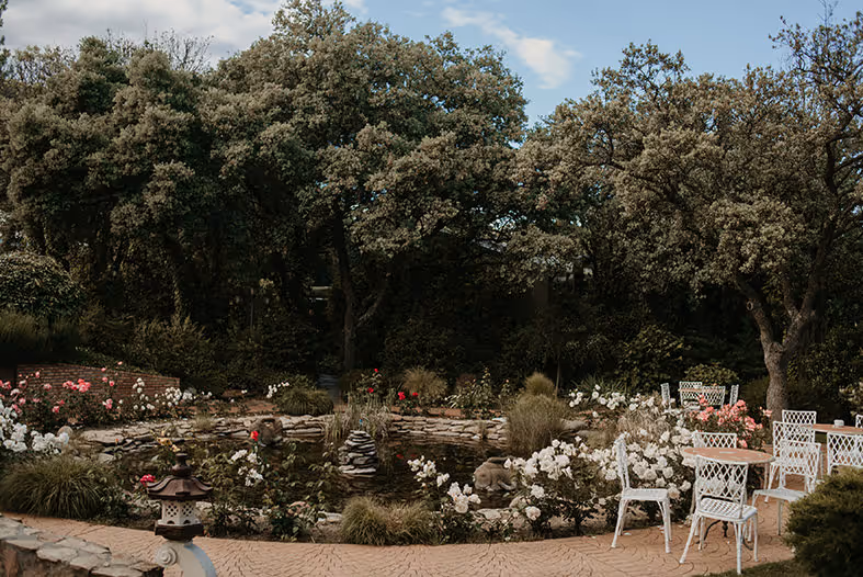 Jardín tranquilo con flores, rocas, sillas blancas y árboles frondosos