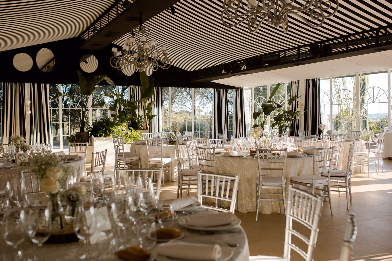 Salón de bodas elegante con mesas blancas, sillas Chiavari y grandes ventanales