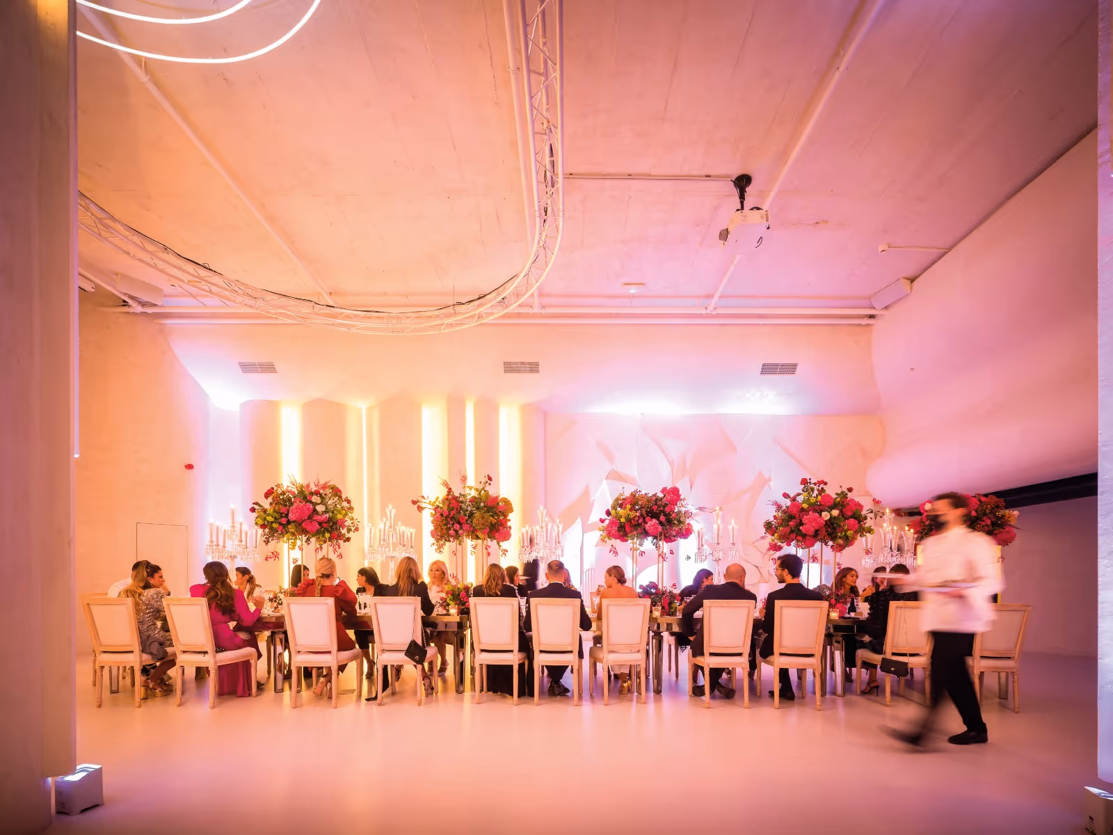 Elegante evento con invitados sentados, iluminación rosa y grandes arreglos florales